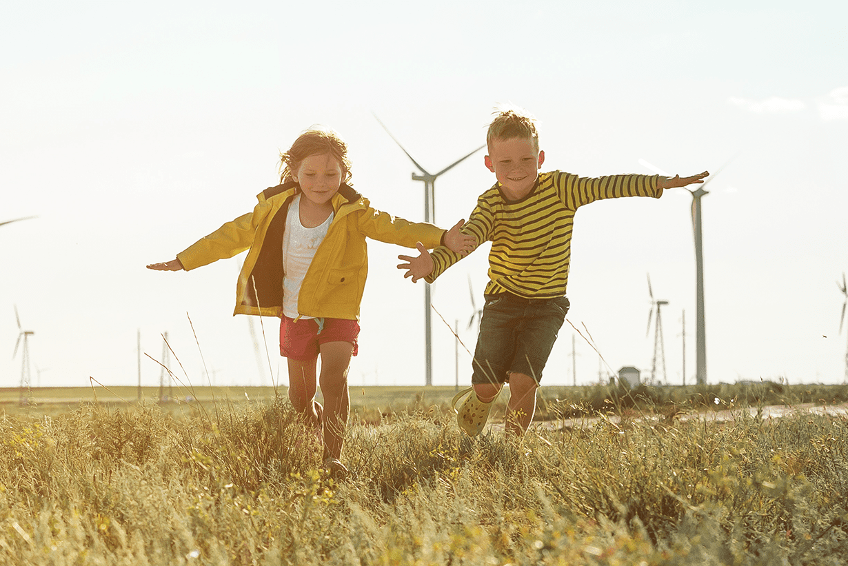 Two children run holding hands with a wind farm in the background