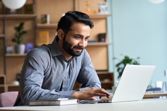Smiling indian business man working on laptop at home office.