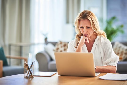 Mature white woman sat at a table using laptop with her hand rested under her chin