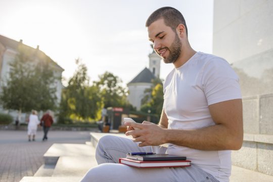 Young adult male sat on a step outside looking down at a mobile phone