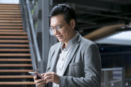 A mature Asian man dressed in business attire looking down at a mobile phone in his hand
