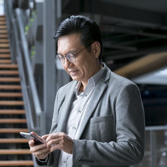 A mature Asian man dressed in business attire looking down at a mobile phone in his hand