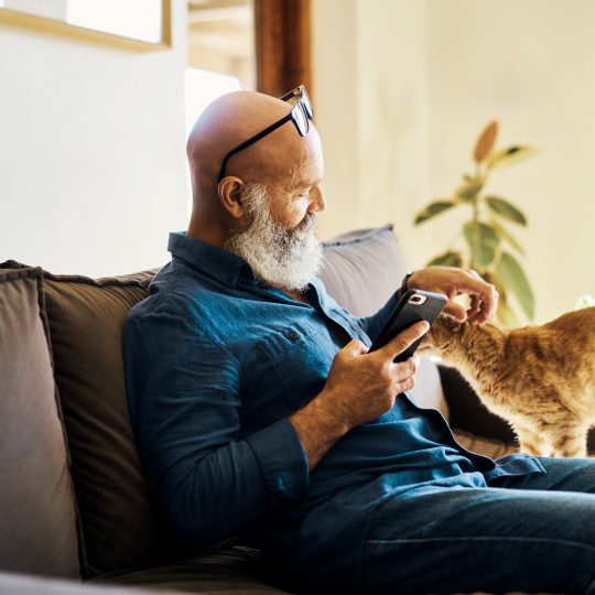 Senior man sat on sofa petting a cat with his left hand, and using a phone in his right hand