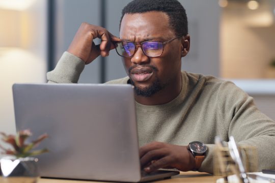 A black adult man holding his glasses looking confused while looking at a computer