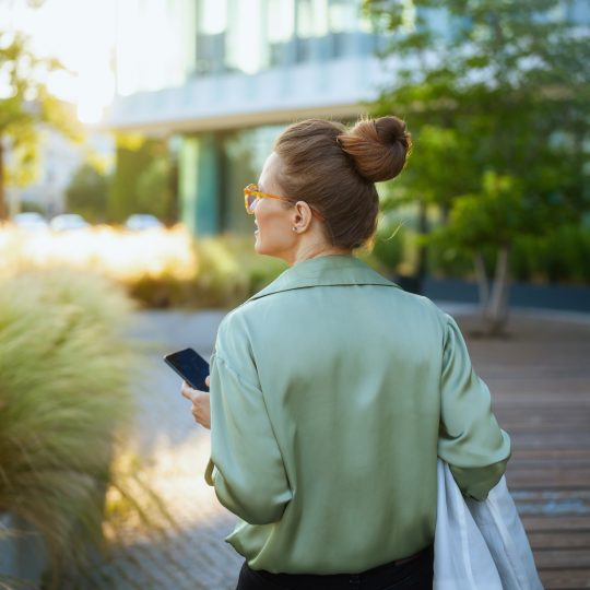 Middle aged white woman faced with her back toward the camera while she walks away holding a mobile phone