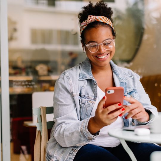 A brown woman sat outside a cafe smiling looking down at a smartphone