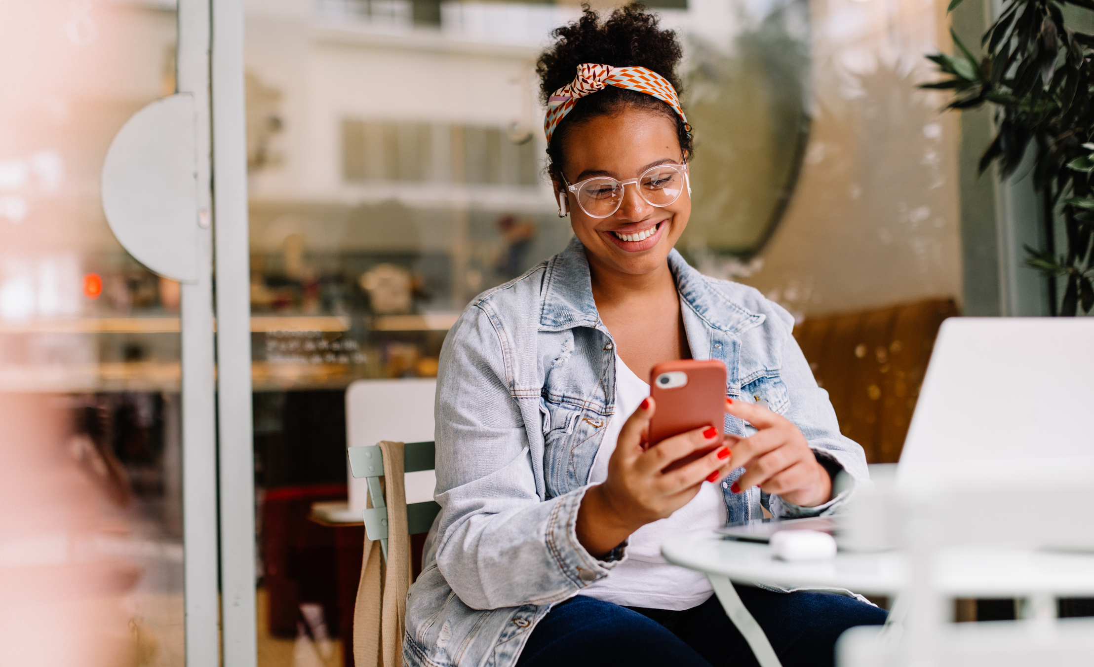 A brown woman sat outside a cafe smiling looking down at a smartphone