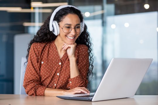 Young beautiful woman in headphones working with laptop inside office at workplace