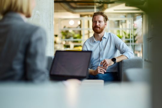 Adult white man sat in a lounge area of an office talking to a female colleague with their body partially visible