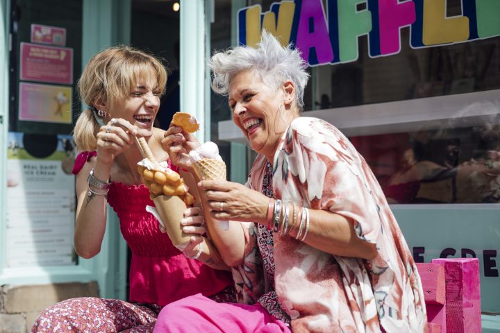 Two women sitting side by side on a bench eating ice cream and waffles together