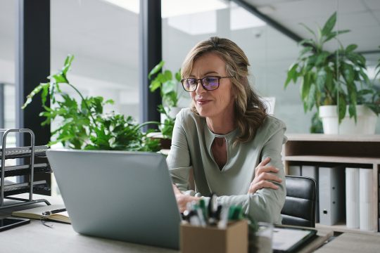 mature business woman at desk in glass office for with arms folded and looking at her laptop