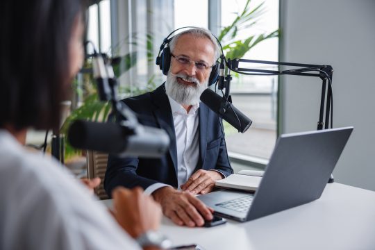 A senior Caucasian male with gray hair and a beard hosts a live podcast
