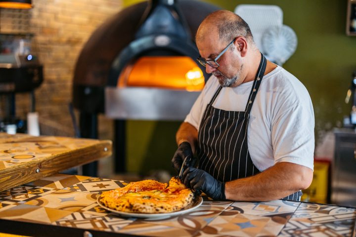 Mature Chef Cutting The Pizza With Scissors In A Commercial Kitchen