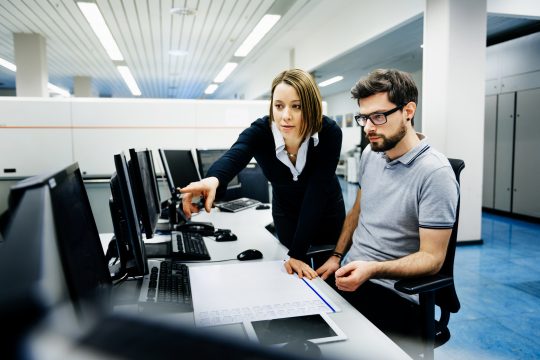 Two IT professionals man and woman in a big control room of a factory talking.