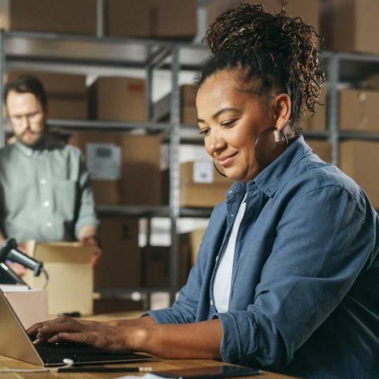Brown woman using laptop in a warehouse while a white man packages a box to her right