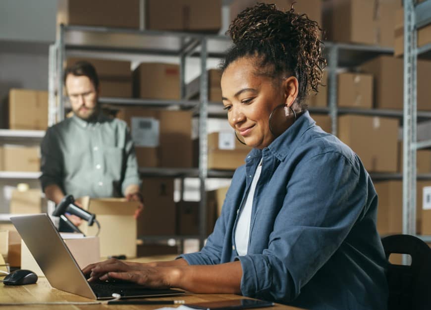 Brown woman using laptop in a warehouse while a white man packages a box to her right