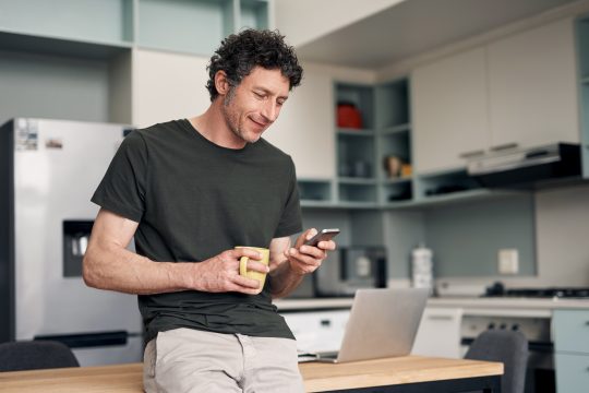 Shot of a mature man using a cellphone while drinking coffee at home