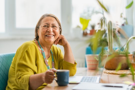 A portrait of a happy fashionable senior woman smiling for the camera while using a laptop at home.