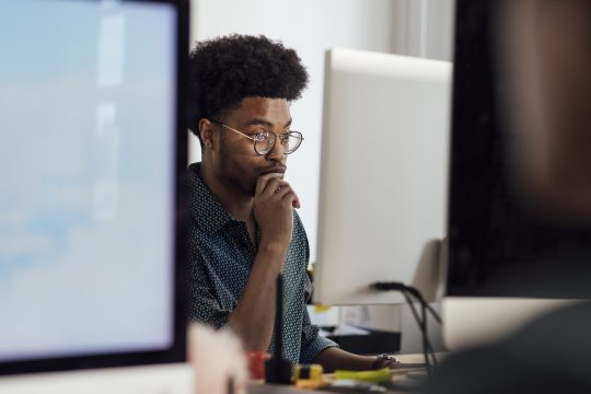 Close-up of a young male office worker sitting at his desk using a computer to complete his work in the office in Newcastle, England. He is wearing smart casual work clothing and he is working to a deadline.