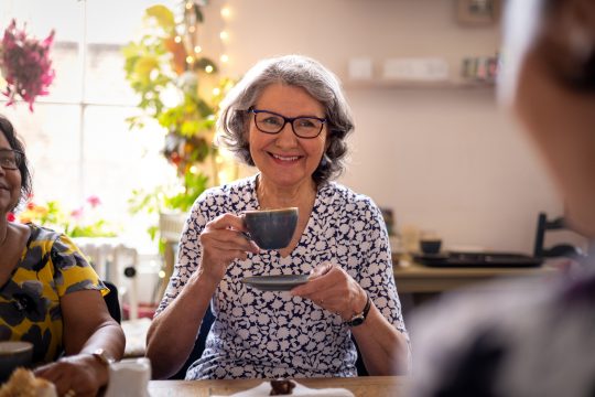 Waist-up over-the-shoulder shot of a mature adult woman talking and smiling with her friends wearing casual clothing in a rustic cafe