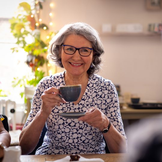 Waist-up over-the-shoulder shot of a mature adult woman talking and smiling with her friends wearing casual clothing in a rustic cafe