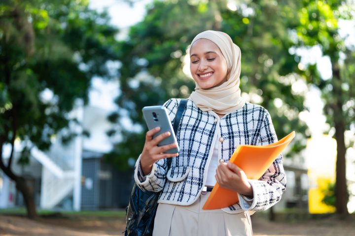 Portrait of young smiling Muslim college girl carrying backpack and using smart phone for online banking