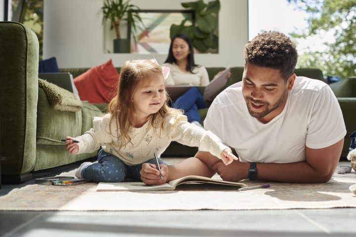 Brown adult man laying on the floor drawing beside a young white toddler