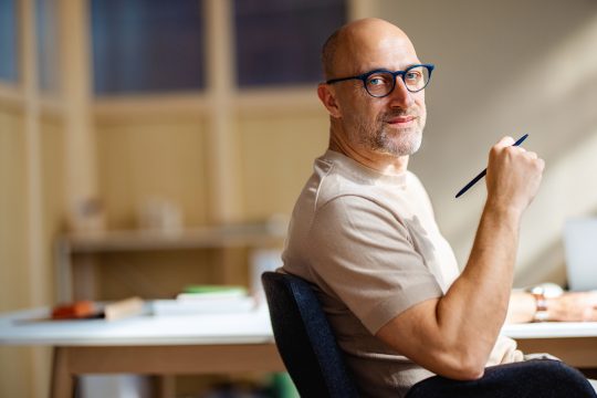 Mature man with glasses working confidently in a modern office setting, holding a pen and looking at the camera.