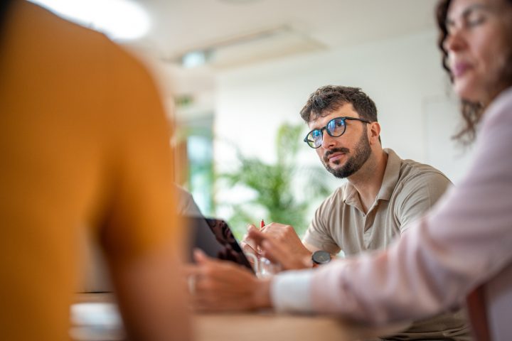 Caucasian male in casual business attire leads a meeting, actively engaging with colleagues of diverse backgrounds in a bright office setting