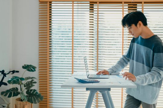 Young Asian man using standing desk at home, working on his laptop and examining financial documents, emphasizing modern work habits and ergonomics. Remotely and finance concept.