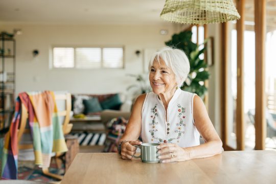 An elderly woman in a white embroidered top smiling while sipping her morning tea