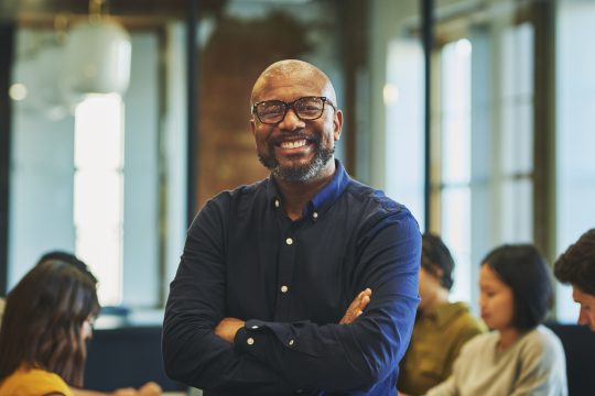 Bearded businessman wearing eyeglasses, crossing arms and smiling cheerfully at camera in office, colleagues meeting in background