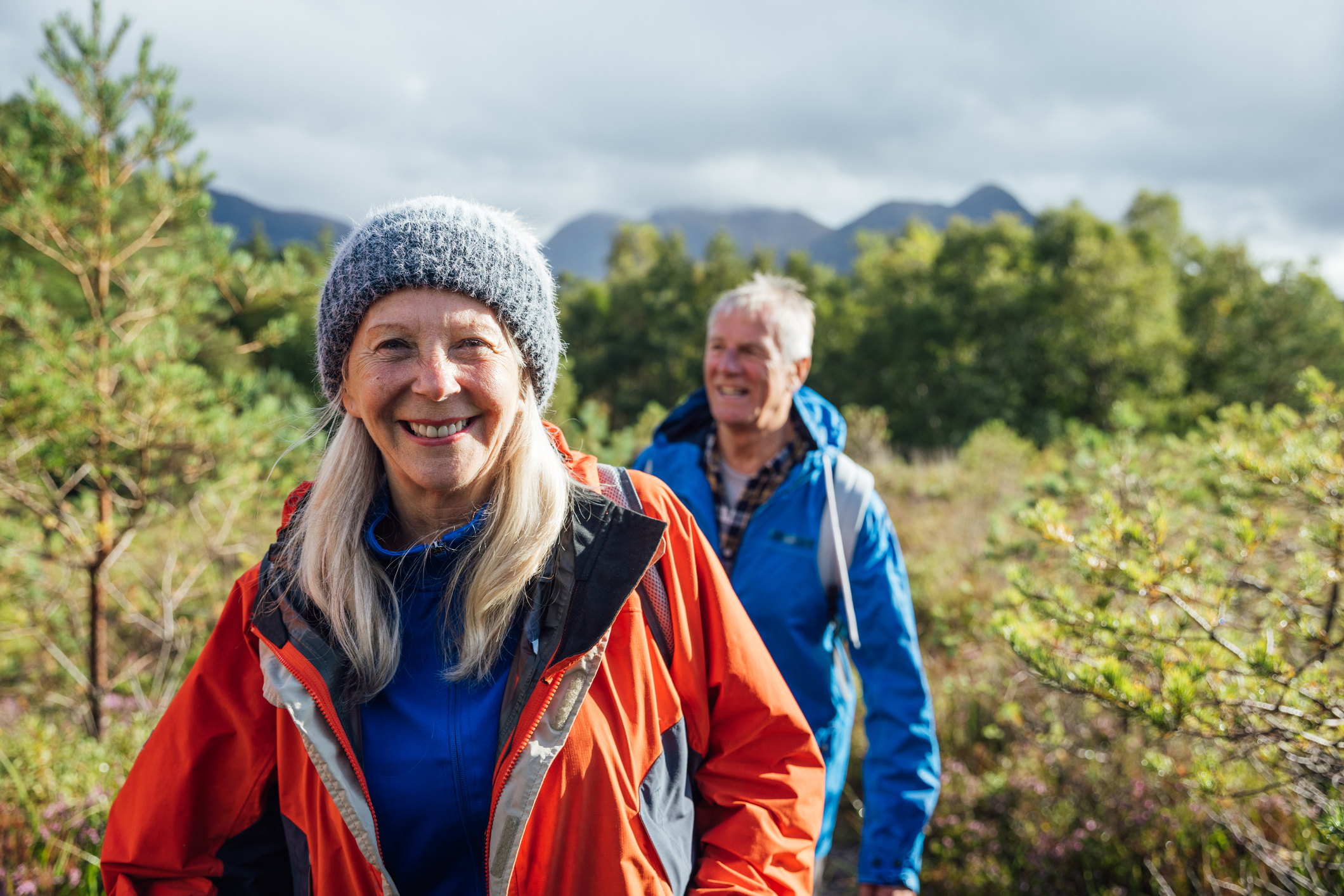 Waist up front view of a senior couple hiking up the mountain landscape surrounding Loch Torridon in the Northwest Highlands, Scotland. The woman is wearing a wooly hat and smiling while looking at the camera.