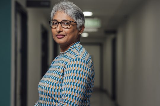 A portrait of a mid adult female doctor, standing in a low lit corridor in a hospital in Newcastle upon Tyne, Proud of Her Work