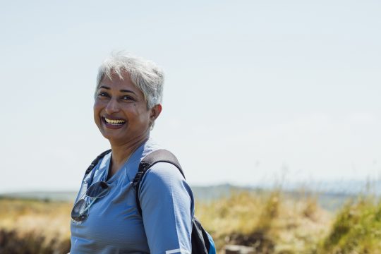 Waist up view of a mature woman hiking on a hill. She is wearing casual sports attire on a summer's day at Hadrian's Wall, Northumberland. She is smiling and looking at the camera. Videos are available similar to this scenario.