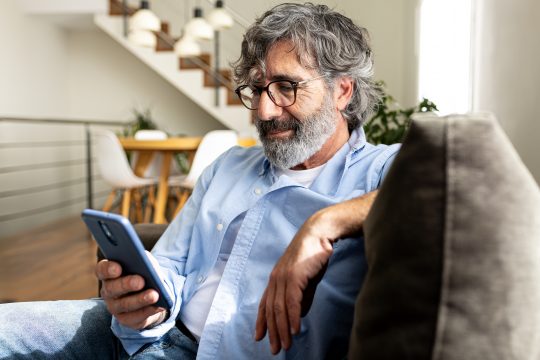 Mature man relaxing at home, sitting on the sofa sending text message with mobile phone. Lifestyle concept.