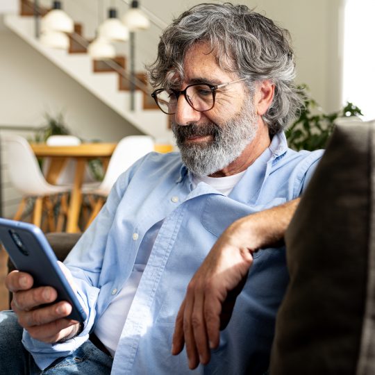 Mature man relaxing at home, sitting on the sofa sending text message with mobile phone. Lifestyle concept.