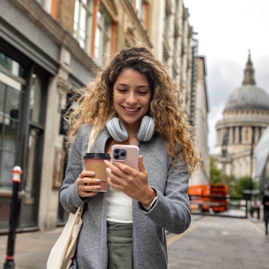 Woman in London using her mobile phone outdoors while drinking a cup of coffee on-the-go